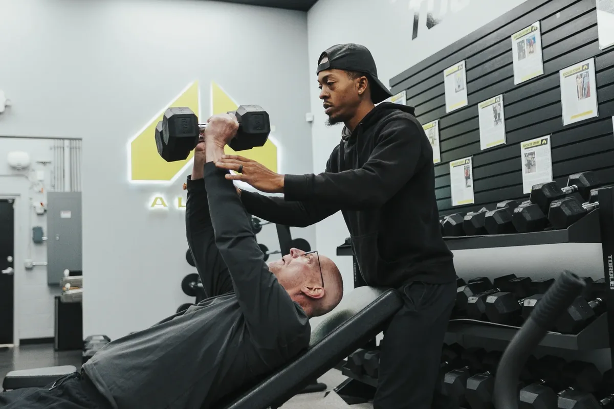 A coach supports an older member's arms as he presses dumbbells overhead from a bench