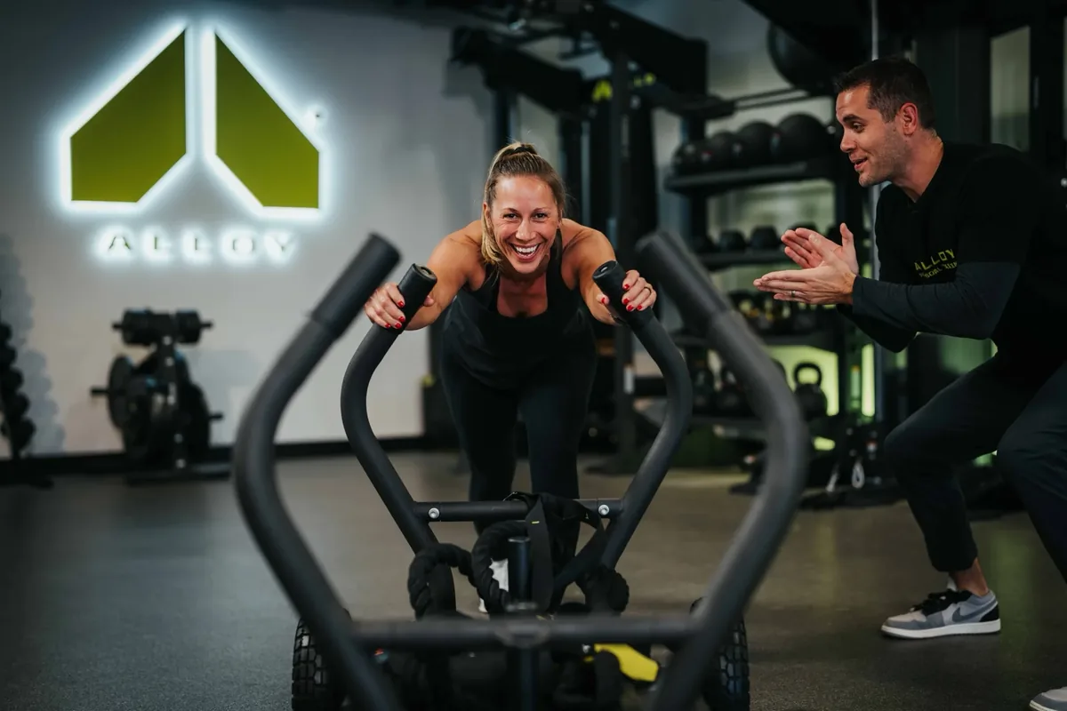 A trainer claps encouragement as a member drives a push sled across the training floor