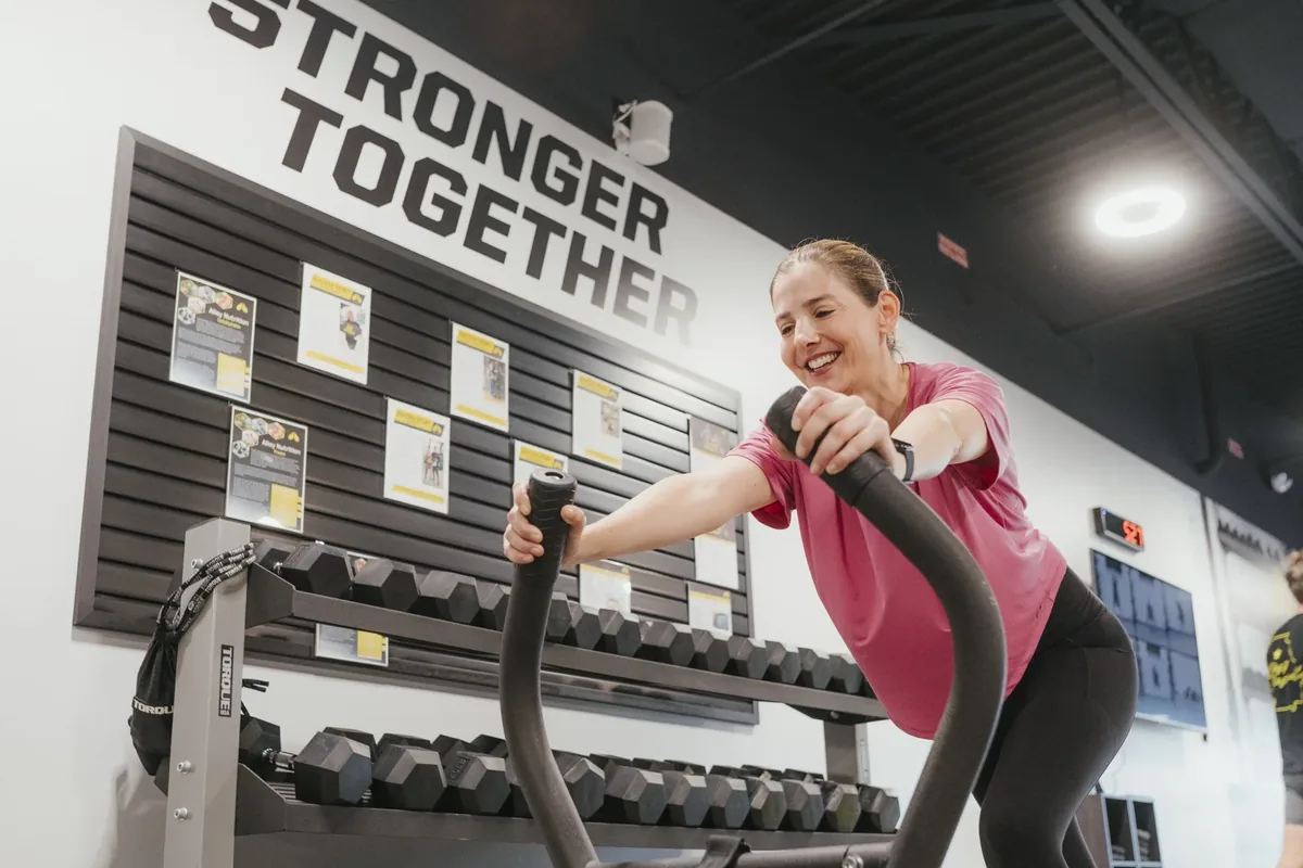 A smiling member works an air bike in front of the dumbbell rack and Stronger Together wall