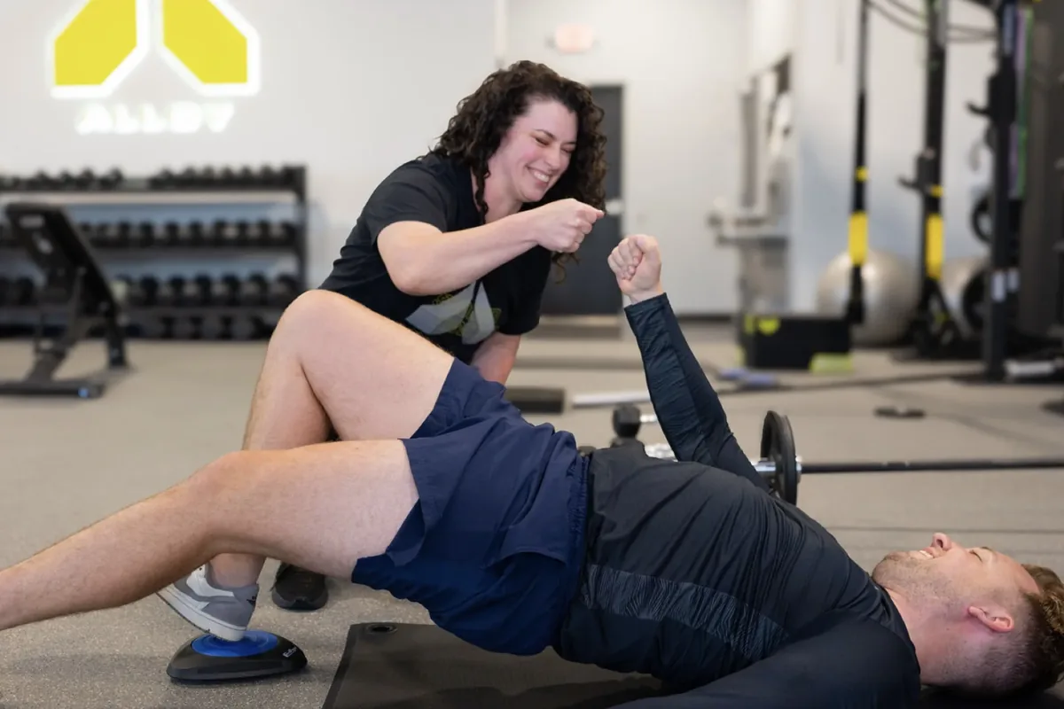 A coach fist-bumps a member holding a single-leg glute bridge with one foot on a glider disc