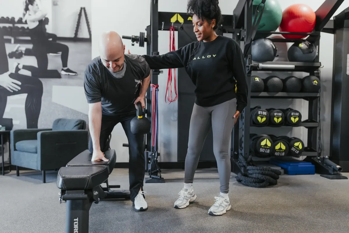 A member rows a kettlebell with one hand braced on a bench while a coach watches beside him