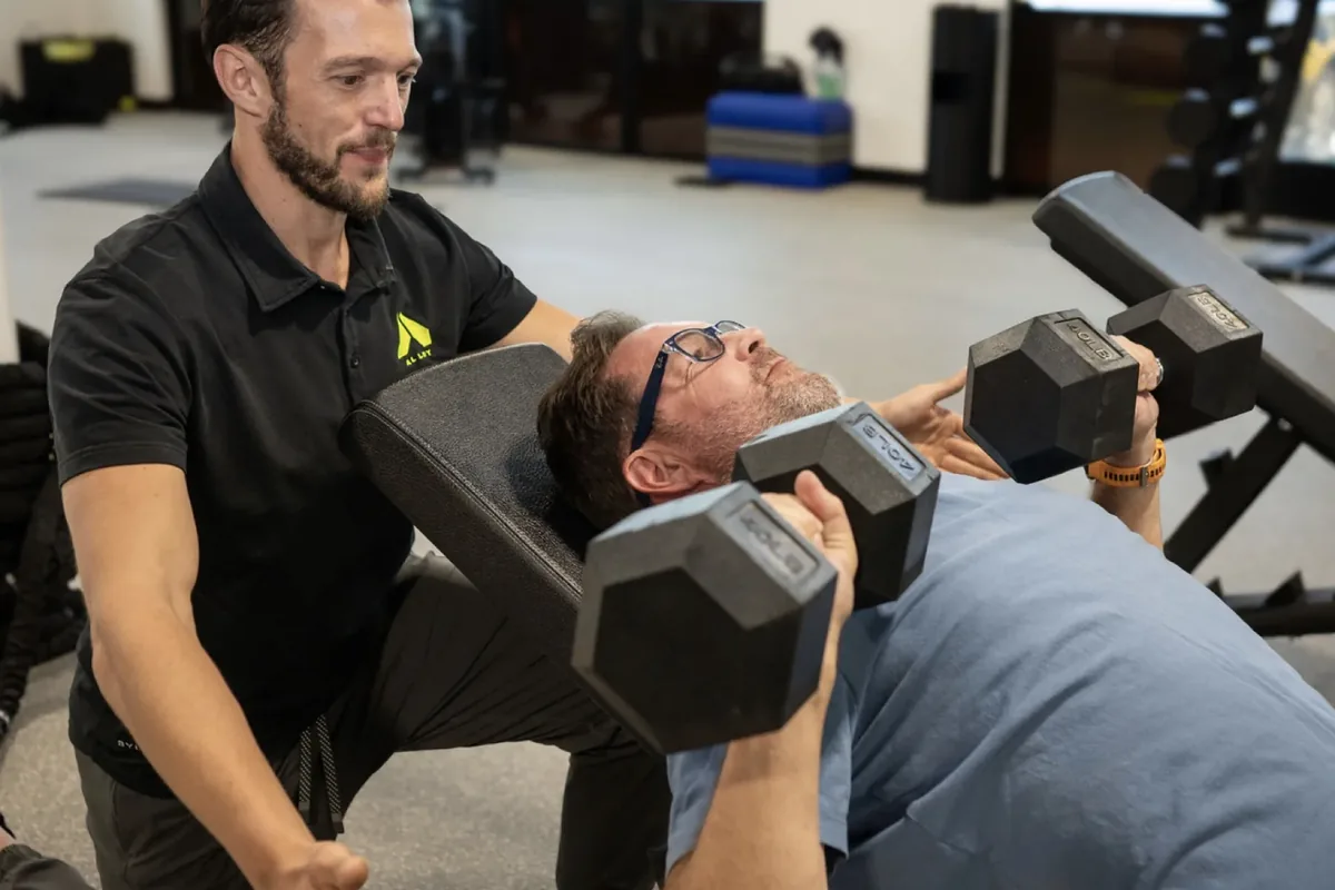 A coach supports the dumbbells as a member presses from an incline bench
