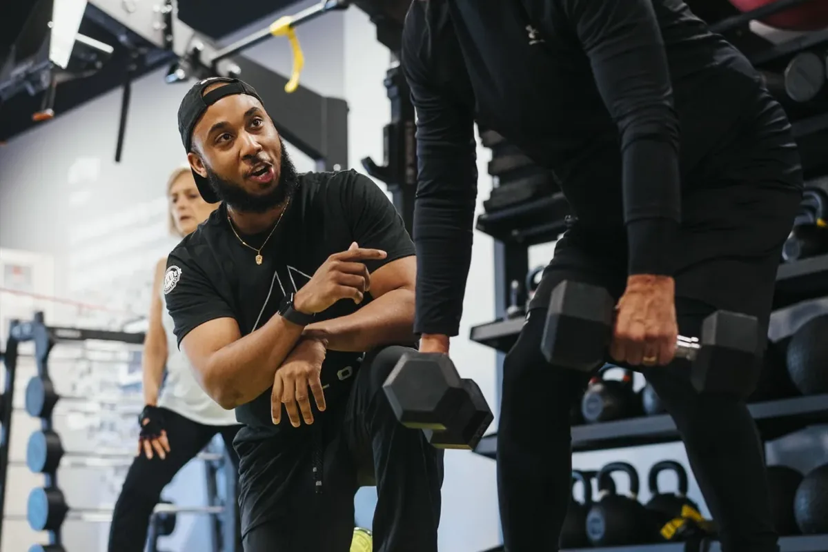 An Alloy coach kneels to guide an older member's dumbbell form