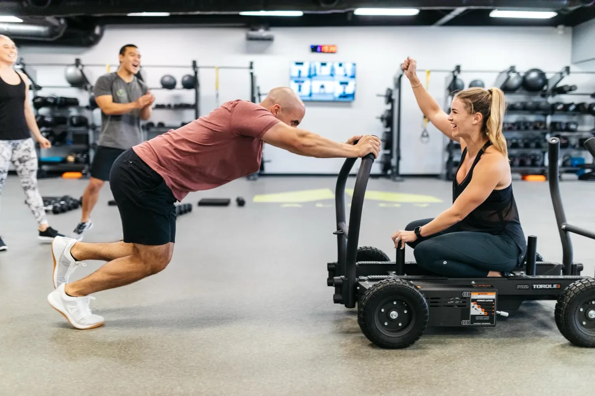Member drives a weighted sled while teammates cheer at Alloy Personal Training — Billings, MT